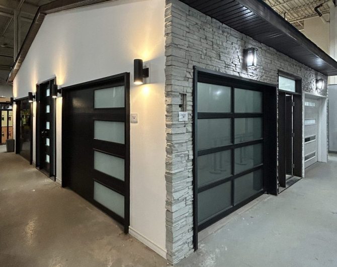 Wide angle view of a door showroom display featuring a white stone facade, black aluminum frame glass garage doors, and exterior lighting.