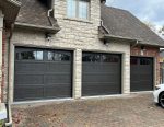 Three dark brown wood-grain texture carriage house garage doors with elegant arched windows installed by Dodds Garage Doors on a luxury light stone and red brick home.