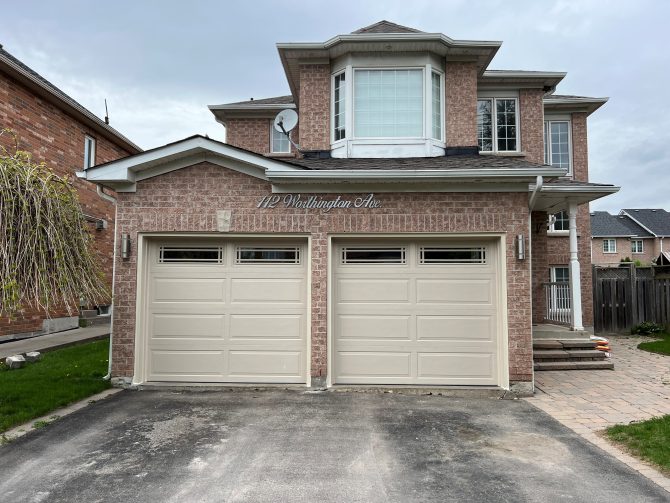 Pair of traditional tan raised-panel garage doors with decorative top windows on a suburban brick home.