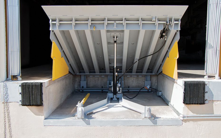 Under-deck view of a heavy-duty mechanical dock leveler showing the lifting mechanism and yellow safety toe guards at a loading bay.