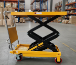 Side view of a yellow manual scissor lift table in a logistics facility, demonstrating the elevated platform height for ergonomic warehouse operations.