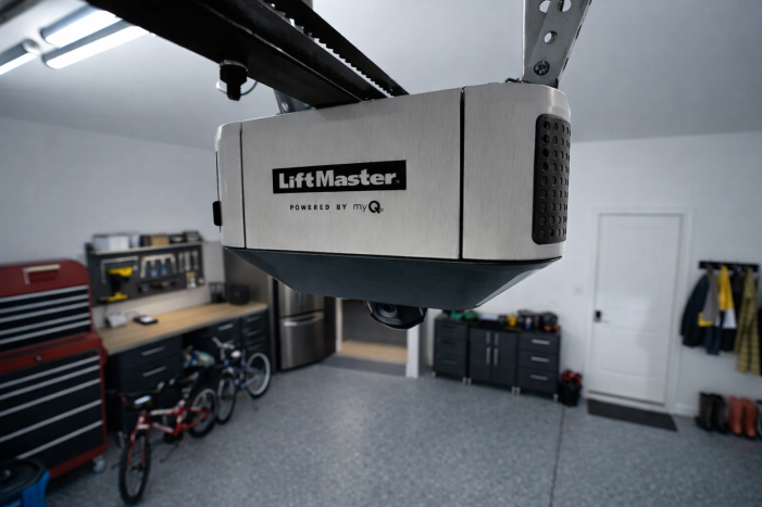 image of a LiftMaster garage door opener with a brushed silver front panel and black perforated side vents, shown from a low angled view against a black background, with the camera lens visible on the underside