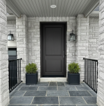 Modern black front entry door with minimalist panels installed on a grey stone house with slate tile porch and black planters.