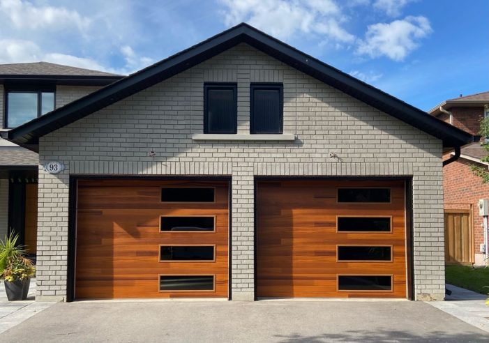 Two modern wood-finish garage doors with horizontal windows installed on a grey brick home.