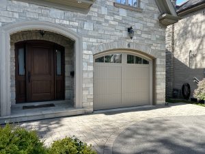 Custom arched garage door in taupe beige installed on a modern stone-veneer home next to a dark wood front entrance by Dodds.