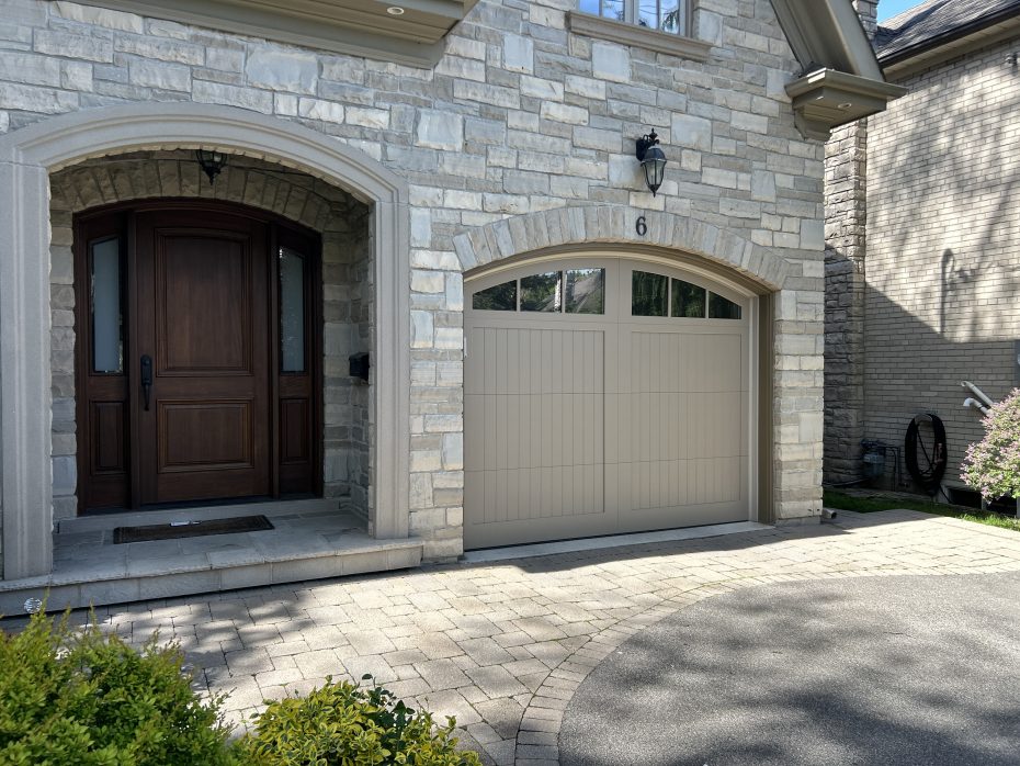 Custom arched garage door in taupe beige installed on a modern stone-veneer home next to a dark wood front entrance by Dodds.