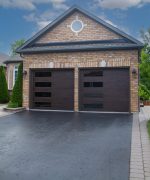 Angled driveway view of two contemporary walnut woodgrain garage doors with left-aligned vertical windows on a brick house.
