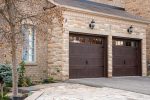 Side view of two long-panel walnut coach house garage doors accenting the textured stone masonry of a modern suburban home.
